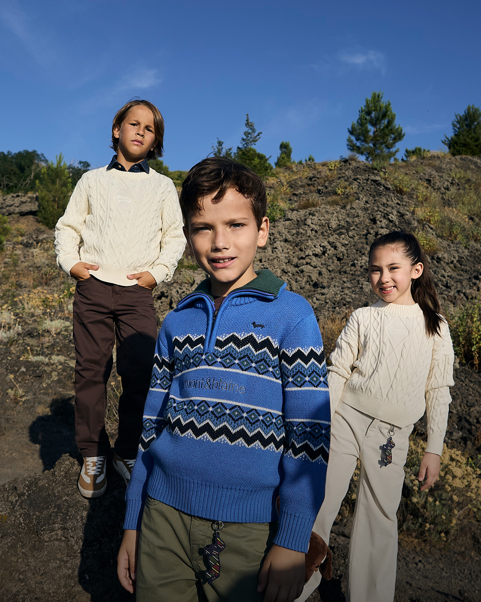 Three children are wearing Harmont&Blaine knitwear items&mdash;in the foreground, a girl in a light cable-knit sweater and a boy in a blue zip-up sweater with dark geometric patterns, while a third child in the background wears a light pullover&mdash;immersed in an outdoor natural setting under a clear blue sky.