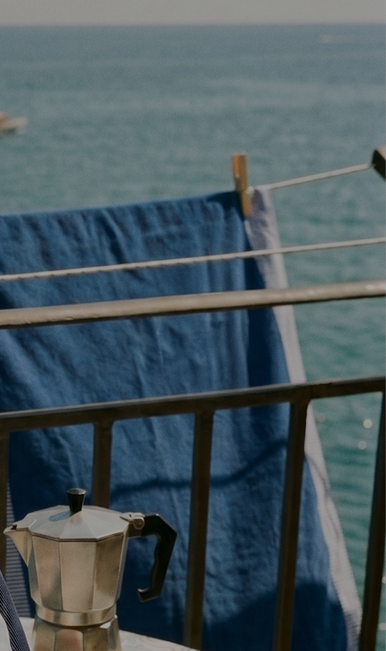 Summer view from a balcony overlooking a calm sea, with a motorboat in the distance. In the foreground, a blue beach towel with white and light blue striped borders is hanging to dry on clotheslines, held by wooden clothespins.
