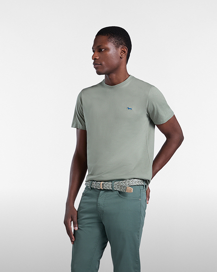 Promotional image focused on the t-shirt category: a man poses in a three-quarter view against a gray background wearing a light sage green short-sleeved t-shirt with a small blue embroidered dachshund logo on the chest. the look is completed with green trousers and a woven belt.