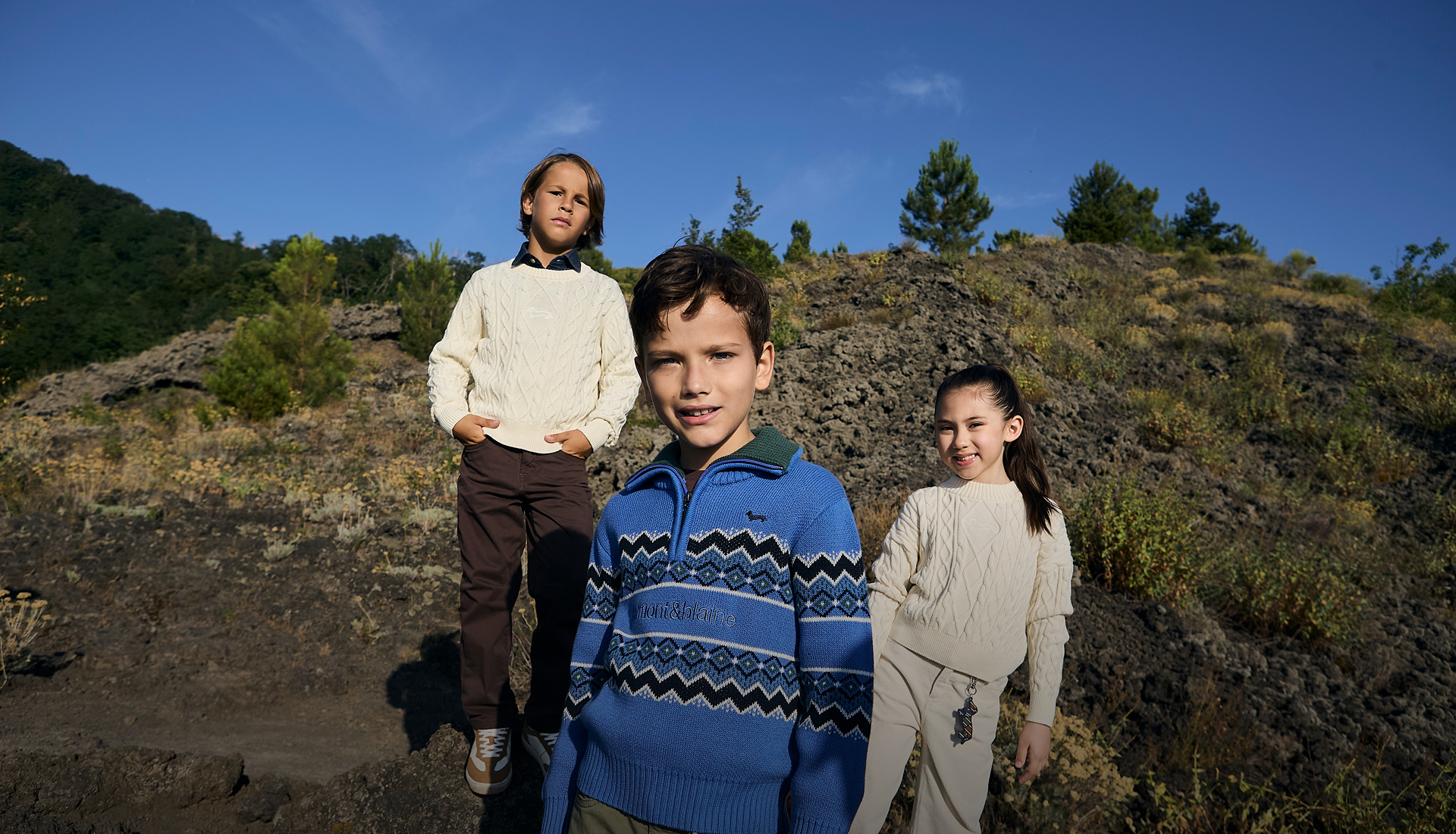 Tre bambini indossano capi di maglieria Harmont & Blaine &mdash; in primo piano una bambina con un pullover chiaro lavorato a trecce e un bambino con un maglione azzurro con zip e motivi geometrici scuri, mentre un terzo bambino sullo sfondo indossa un pullover chiaro &mdash; immersi in un contesto naturale all'aperto sotto un cielo azzurro.