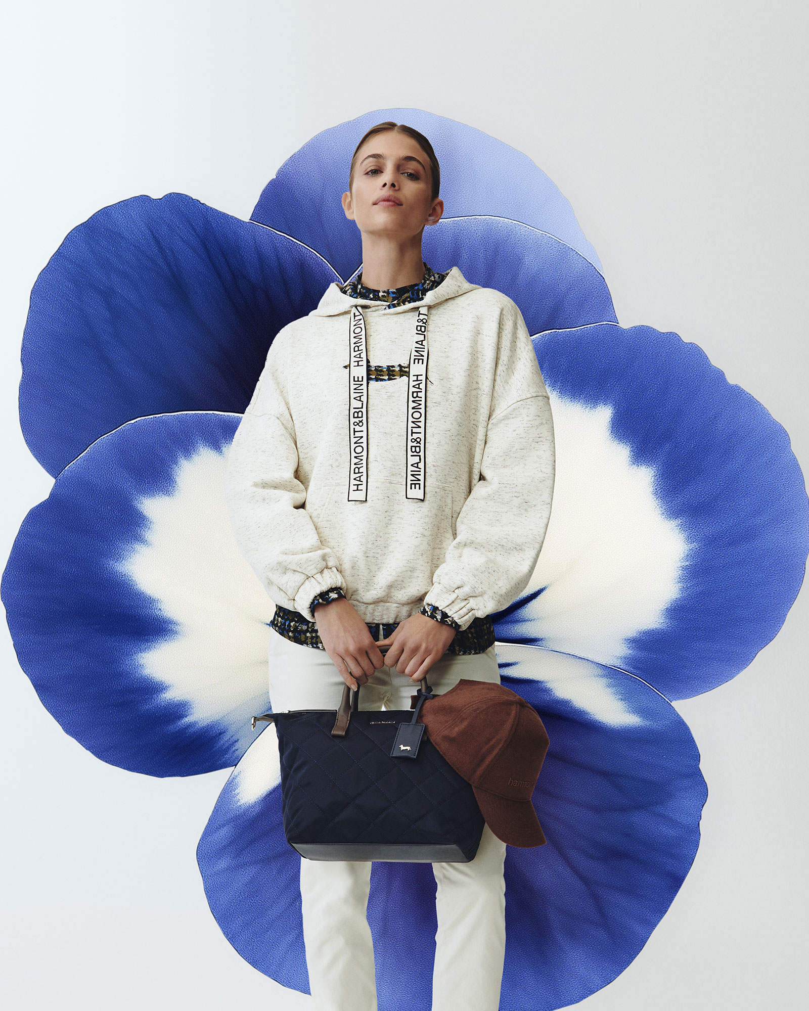 Donna con felpa chiara con lacci logati, pantaloni chiari e borsa trapuntata blu con cappellino marrone