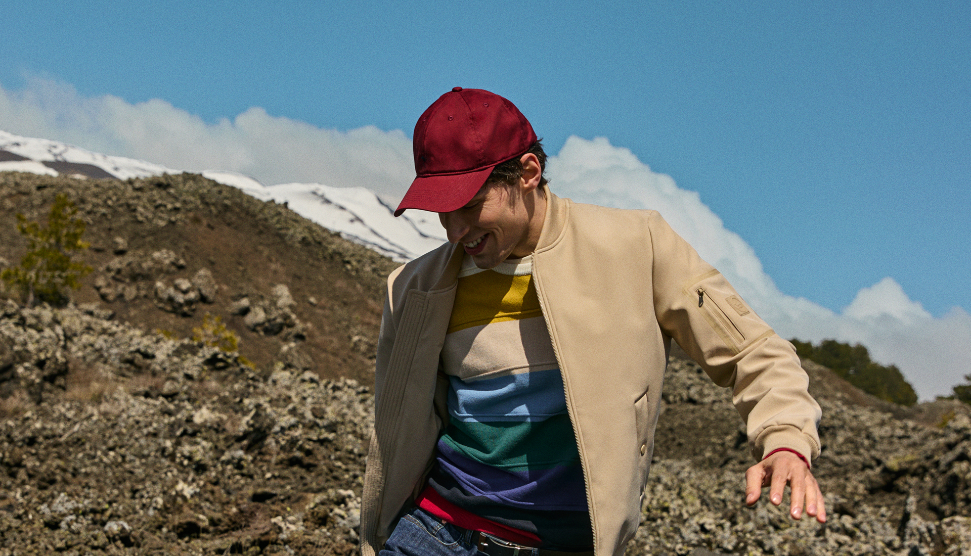  An image of a smiling man outdoors with a mountainous and volcanic landscape in the background. The man wears a burgundy baseball cap, an open beige bomber jacket, and a striped colorful sweater. He is looking downwards with an amused expression, in motion. The sky is blue with white clouds.