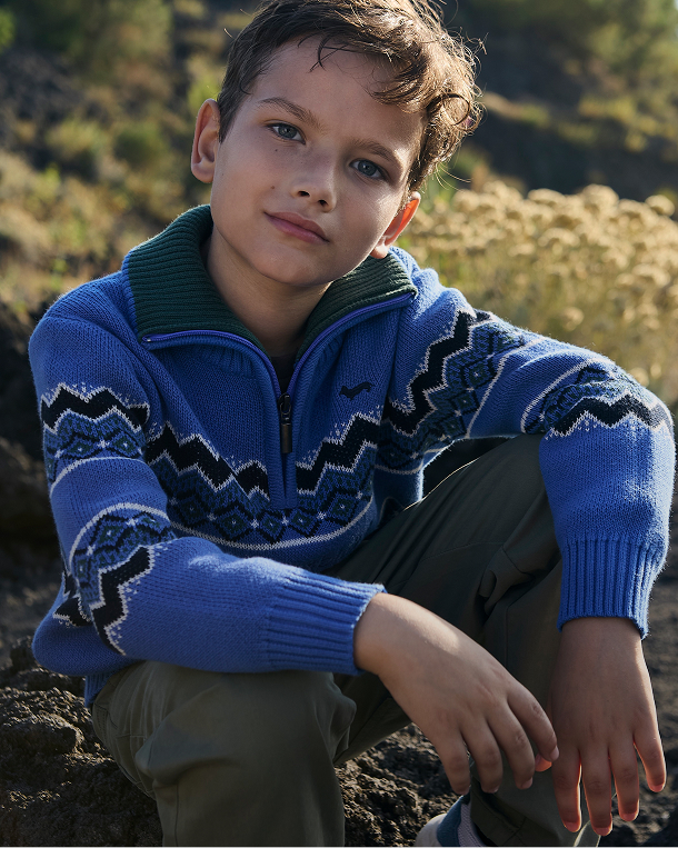 A boy portrayed outdoors looking straight ahead, seated against a rocky and earthy slope covered with dry vegetation, wearing a vivid blue zip-up high-neck sweater featuring a geometric zig-zag striped pattern in dark and light tones, a dark green collar, and olive green cargo pants.
