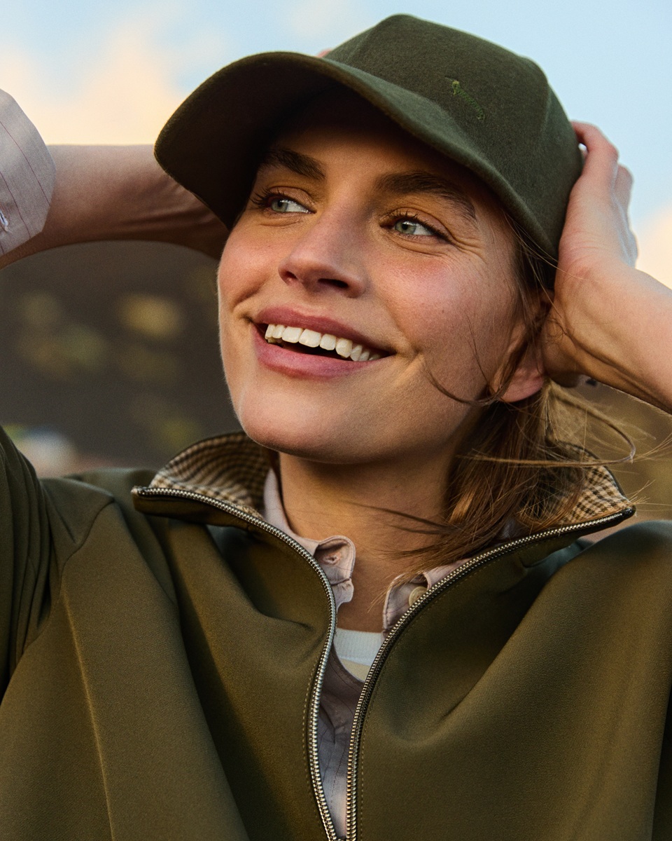 Close-up of a smiling female model wearing an olive green baseball cap with a logo and a matching zipped sweatshirt.
