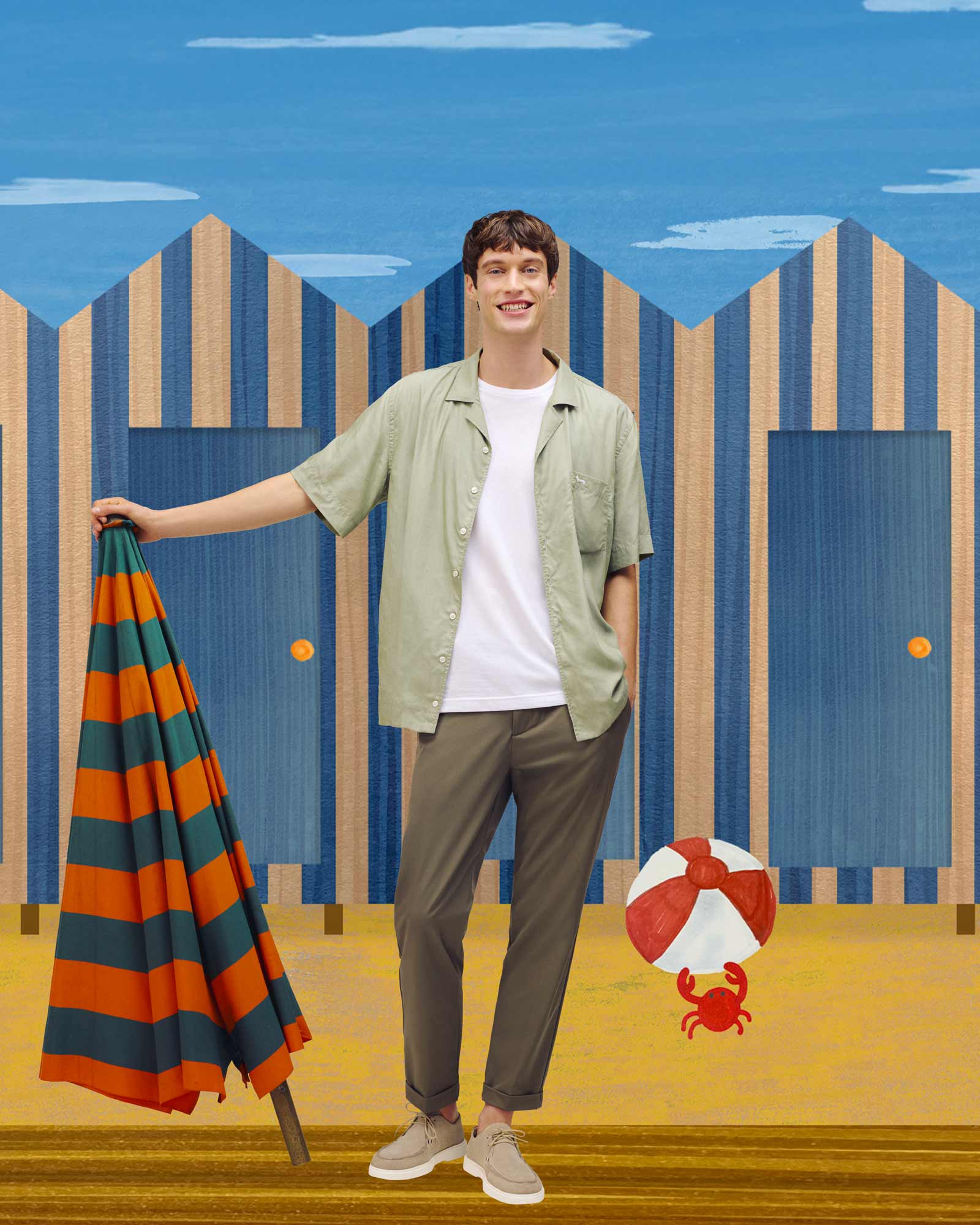 Smiling man in a light green short-sleeve shirt over a white T-shirt, standing in front of striped beach cabins and holding a striped towel — relaxed summer style.