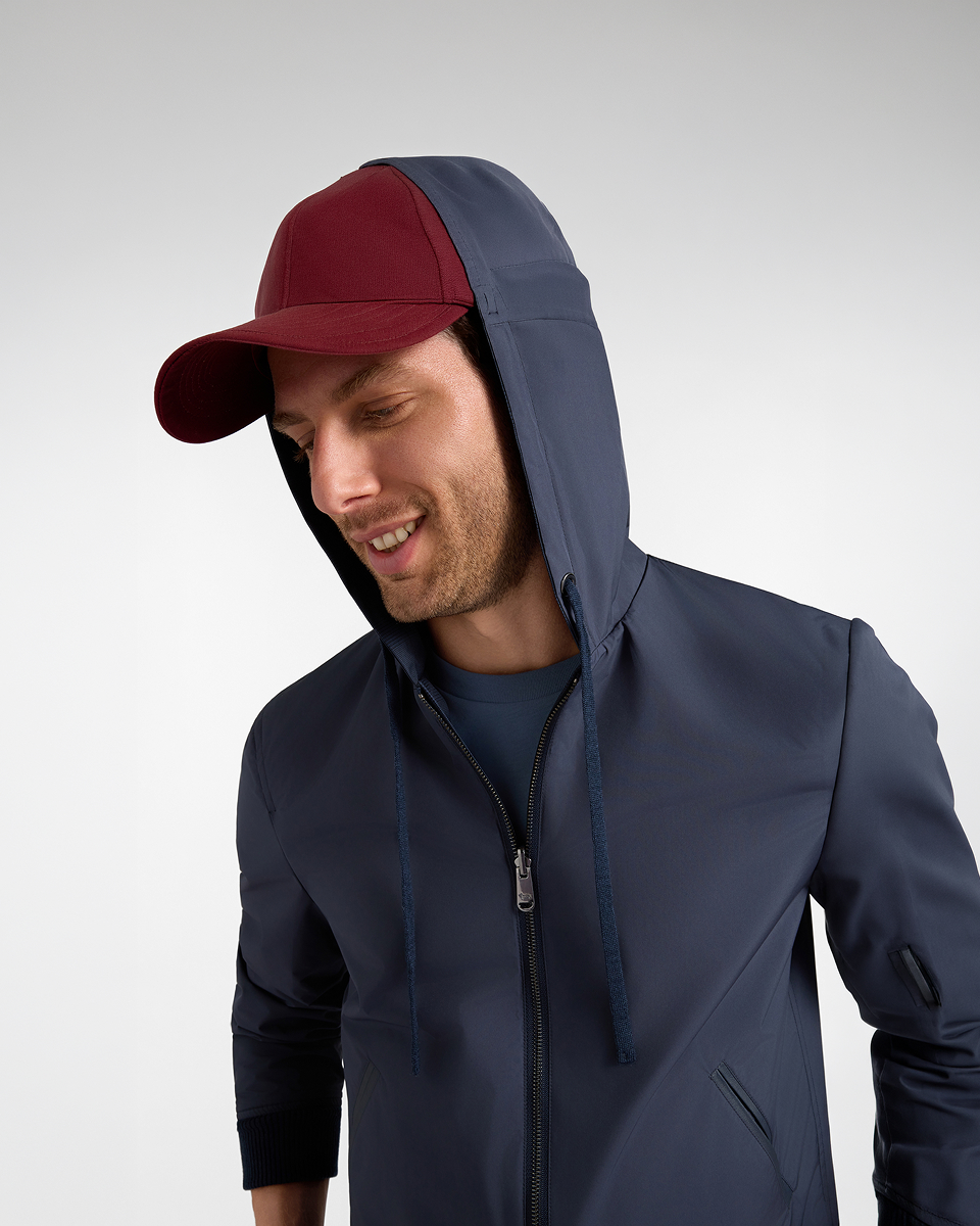 Smiling man wearing a dark blue hooded jacket with a burgundy baseball cap styled underneath the hood.