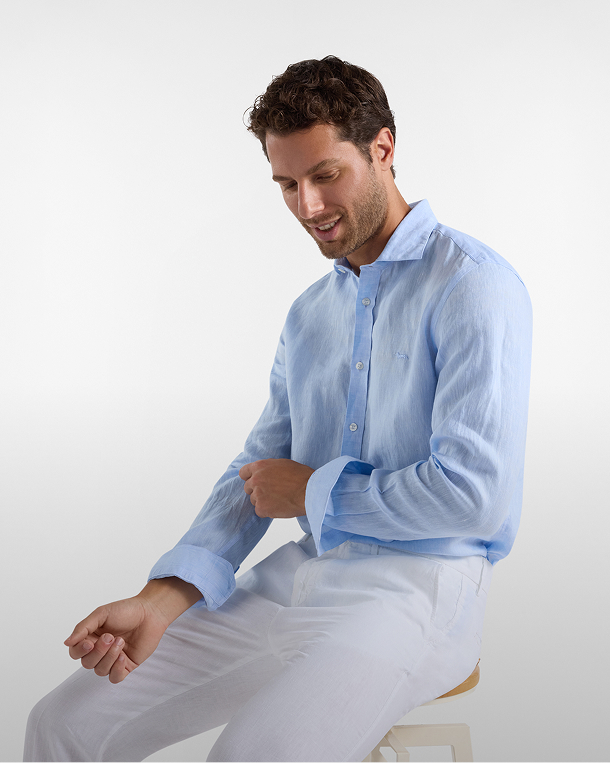uomo in camicia di lino azzurra e pantaloni bianchi &egrave; seduto, guarda in basso sorridendo e si sistema la manica. Lo sfondo &egrave; chiaro e sfumato. In basso compare il testo nuovi arrivi.