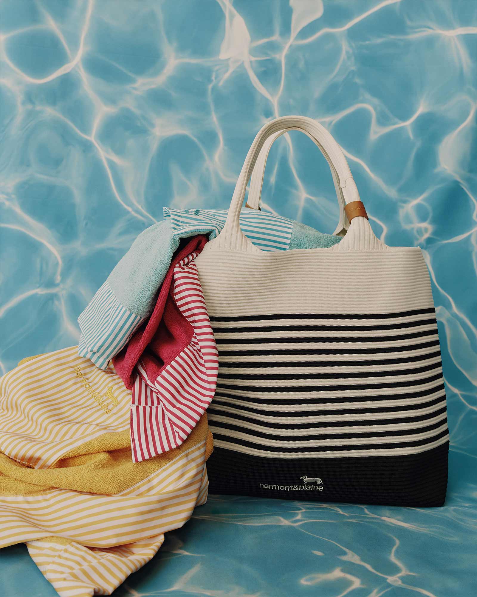 Bolsa de playa a rayas blancas y negras con prendas coloridas de Harmont & Blaine sobre un fondo azul que simula agua.