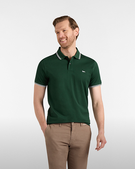 Promotional image focused on the polo category: A smiling man poses against a light gray background wearing a dark green short-sleeved polo shirt. The polo features a classic collar and sleeve cuffs detailed with contrasting thin white stripes, along with the dachshund logo embroidered on the left chest. He is also wearing light beige trousers.