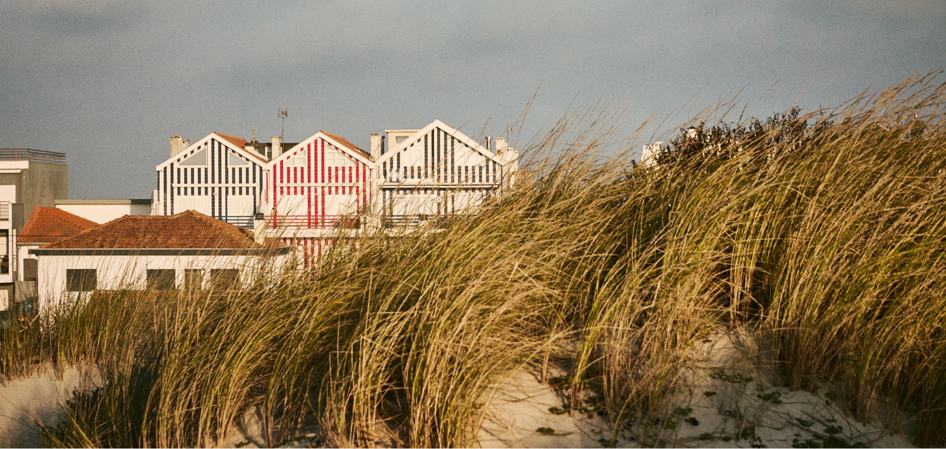 Typical houses with vertical colored stripes emerging behind a coastal dune covered in tall, golden grass, under a cloudy sky. Summer essentials FOR HER. Lightness and style, every day.