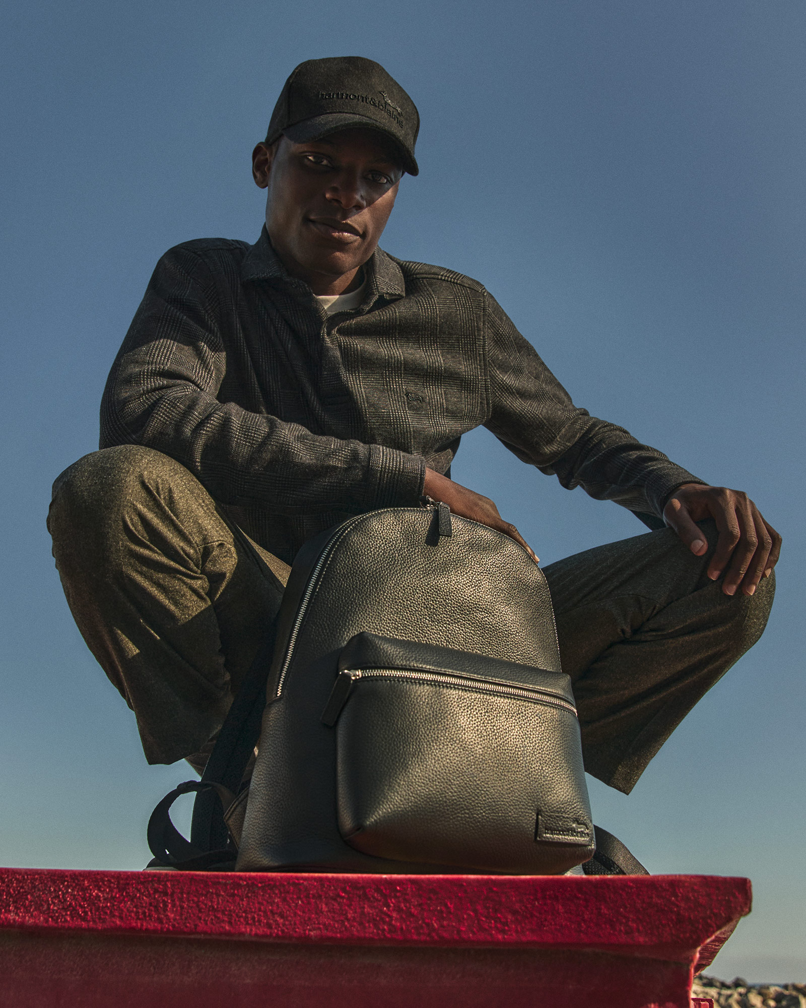 Man wearing a dark shirt and black cap holding a black leather backpack.