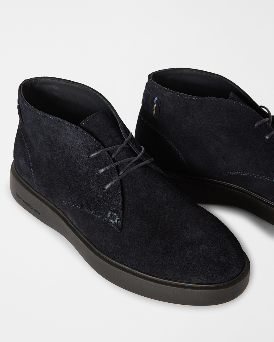A close-up shot on a light background showing a pair of men's dark blue suede chukka boots with matching laces, visible stitching, and a thick dark rubber sole.