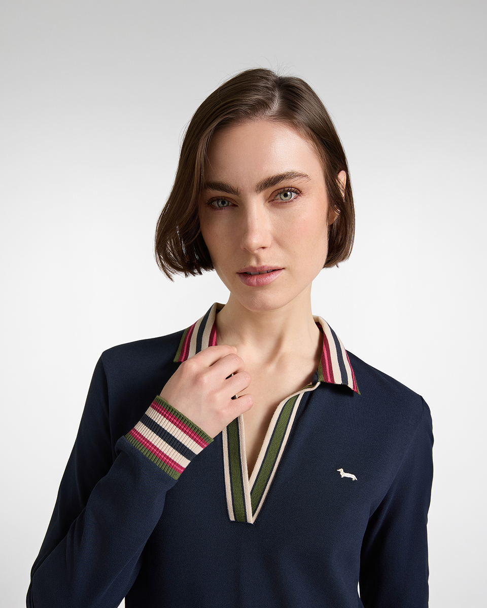 A close-up shot of a woman with short brown hair wearing a dark blue long-sleeved polo shirt featuring a V-neckline, multicoloured striped trim on the collar and cuffs, and a small embroidered logo on the chest.