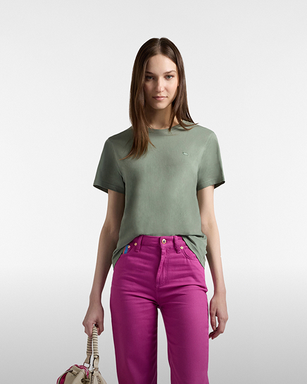 Promotional image focused on the women's t-shirt category: a model poses facing forward against a light gray gradient background. she wears a sage green short-sleeved t-shirt with a small white embroidered dachshund logo on the chest. the look is completed with bright fuchsia high-waisted trousers. in her right hand, she holds a neutral-colored handbag. her hair is long and brown.