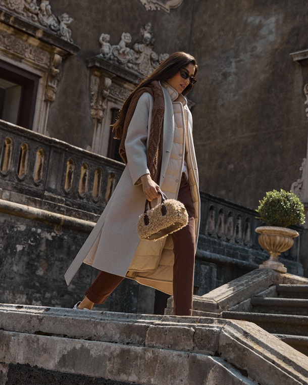 An editorial shot for a women's outerwear focus, featuring a model descending a stone staircase against a historic building backdrop. She wears a sophisticated long, ivory-colored double wool coat, structured yet flowing, over a coordinating quilted lining. The look is completed with brown cigarette trousers, sunglasses, and a textured shearling handbag.