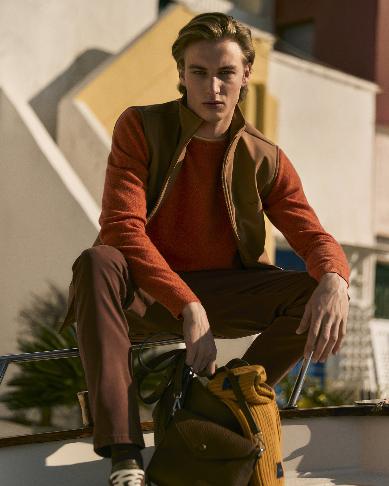 Uomo con maglia arancione e gilet marrone, pantaloni marroni e borsa gialla