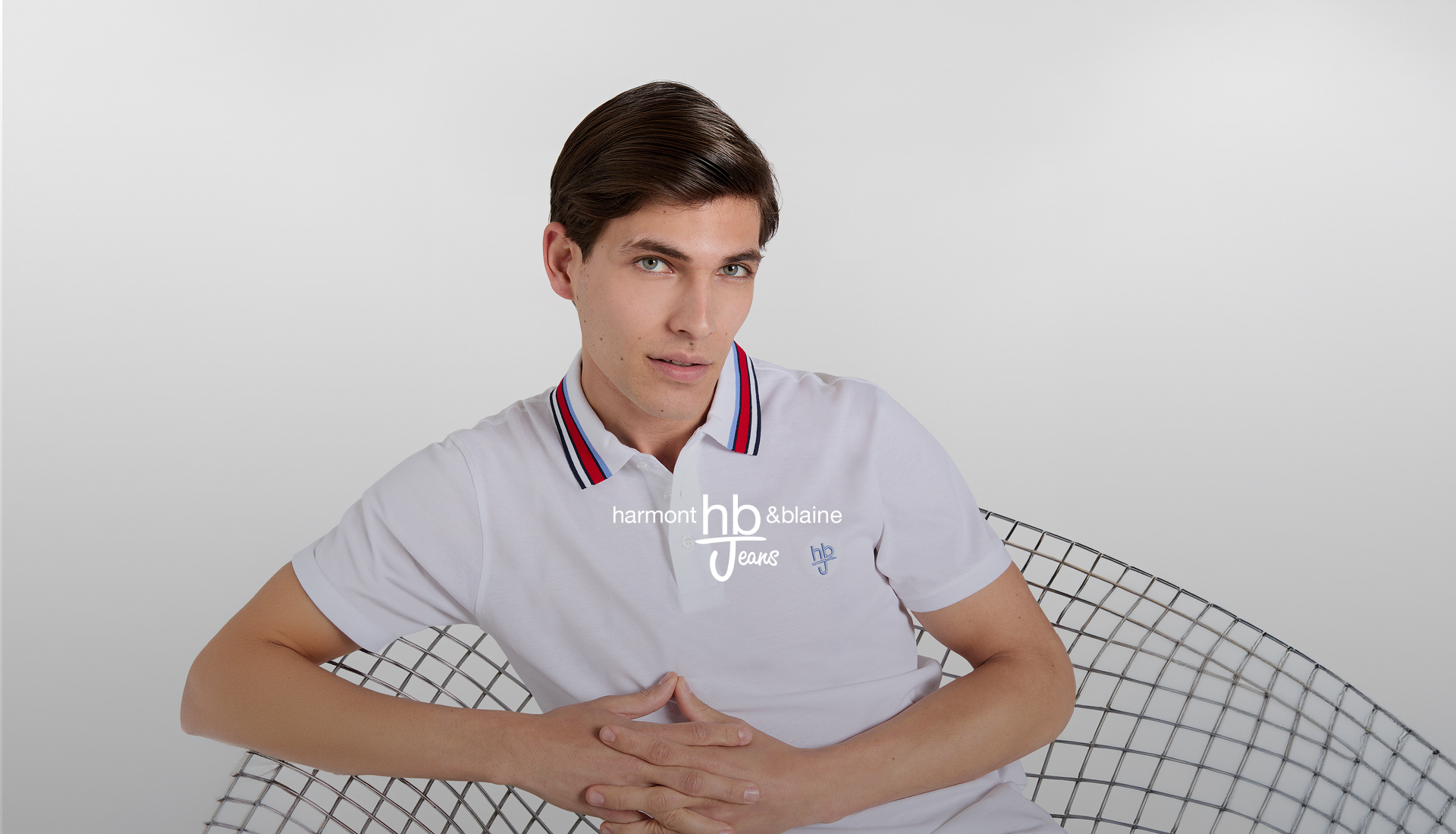 Modello seduto su una sedia a rete metallica che indossa una polo bianca con colletto a righe rosse, blu e bianche. In sovrimpressione il logo "harmont & blaine hb Jeans" e il testo promozionale: "-30% su Tutto, -40% su Giacche e Giubbotti.