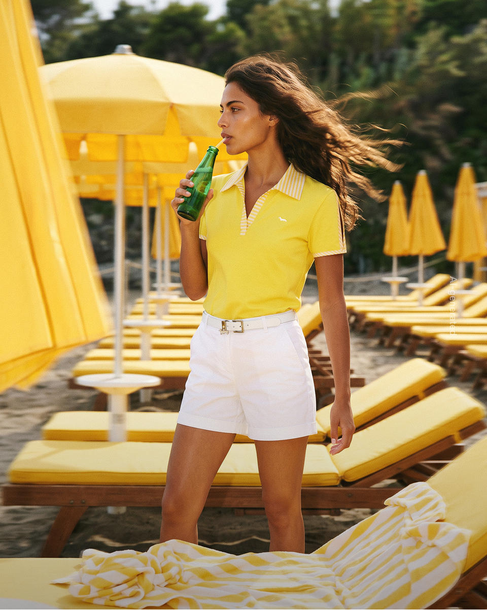 Promotional image focused on the women's polo category (generated with artificial intelligence): a model stands on a beach surrounded by yellow umbrellas and sunbeds, drinking through a straw from a green glass bottle. she wears a bright yellow short-sleeved polo shirt featuring a collar and trim with yellow and white stripes, and a white embroidered dachshund logo on the chest. the look is completed with white bermuda shorts and a matching belt.