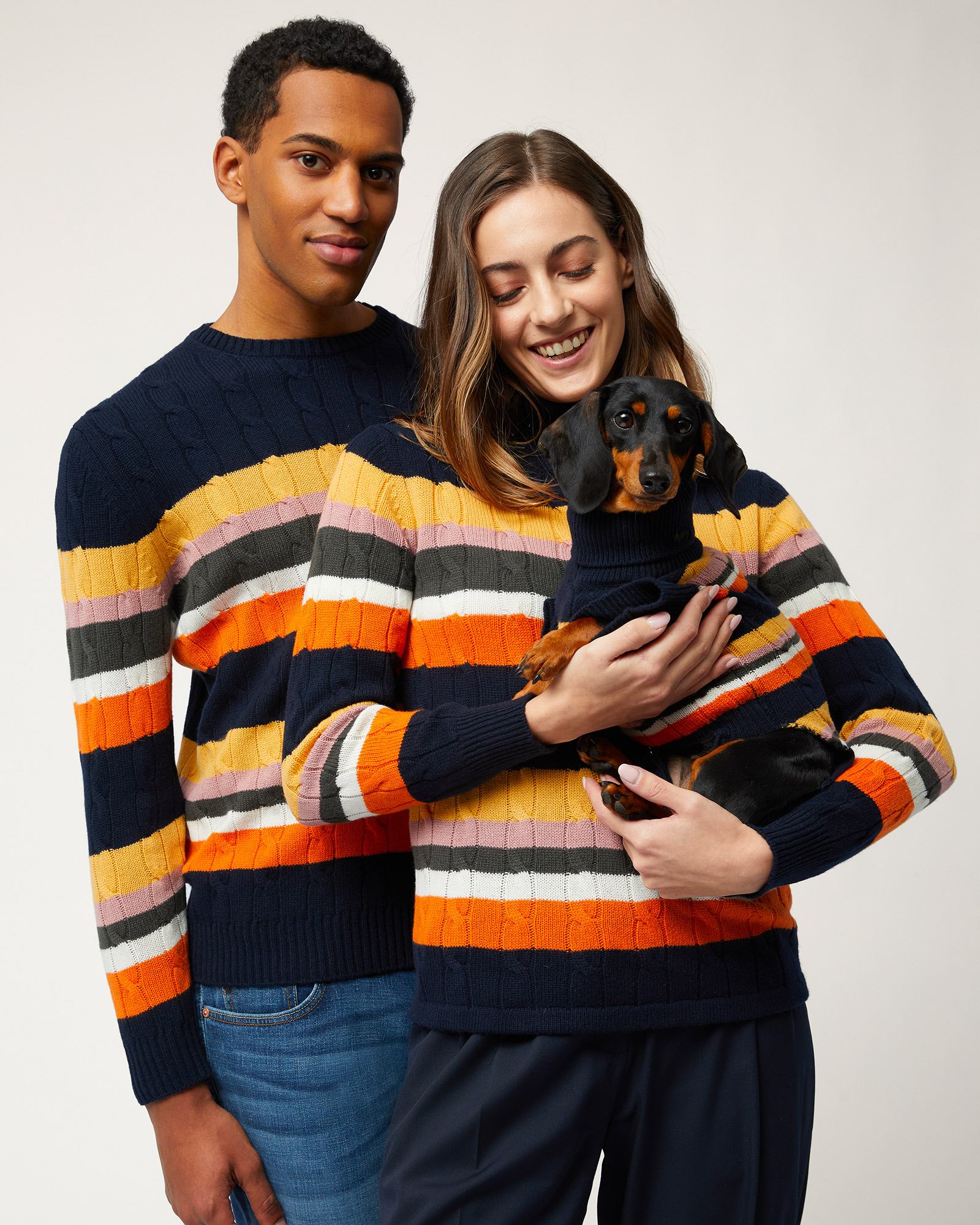 Pareja con suéteres de rayas de colores y un perro salchicha con suéter a juego.