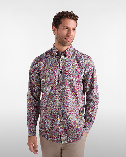 A man with curly brown hair and a short beard wearing a long-sleeved shirt with a multicoloured paisley pattern in shades of purple, pink, and green, paired with beige trousers.