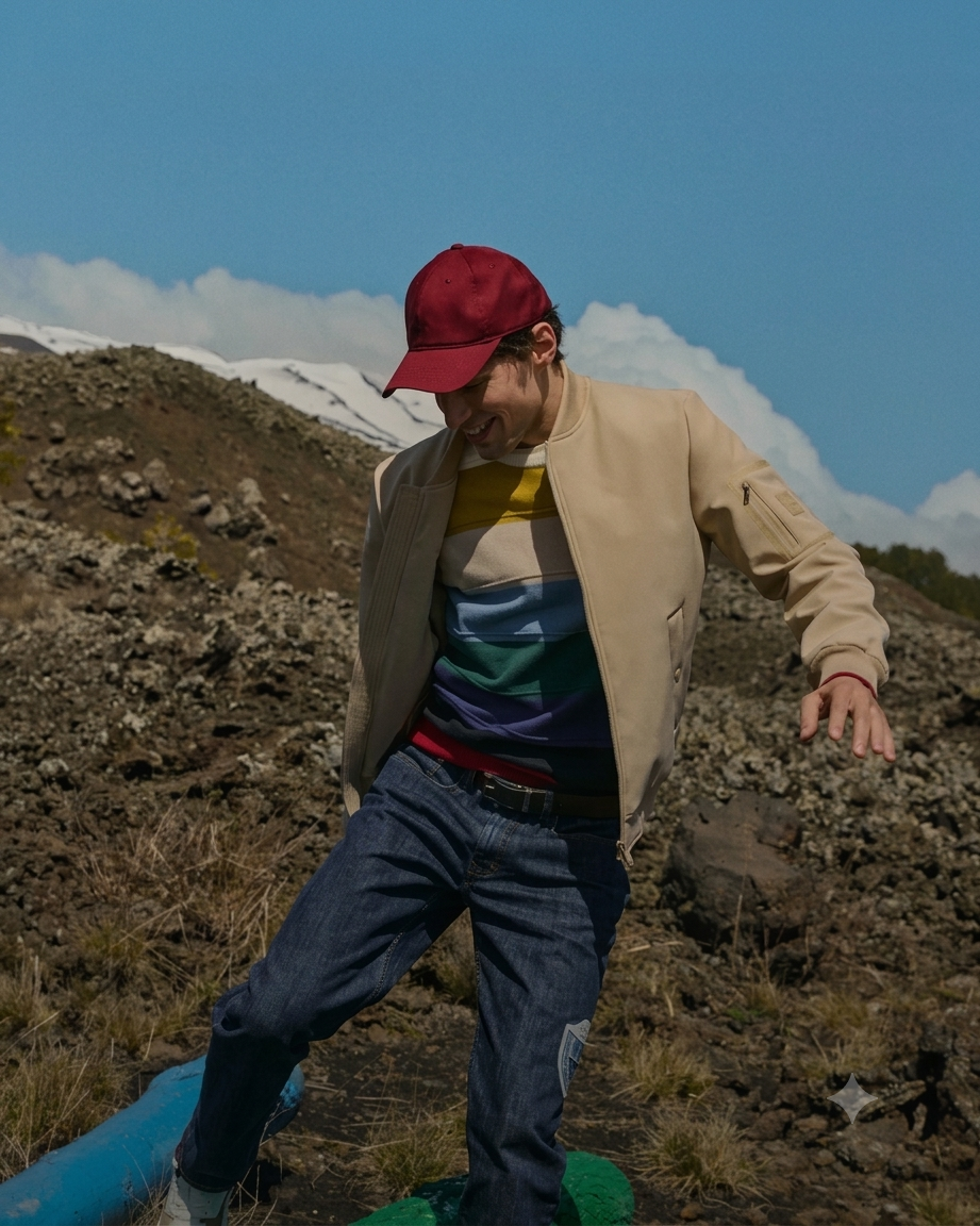 In uno scenario vulcanico e montano sotto un cielo limpido, un uomo si muove con spensieratezza vestendo la collezione preview Uomo Autunno/Inverno 2026. Sorridente e catturato in un gesto dinamico, indossa un cappellino rosso bordeaux, un bomber aperto color sabbia e una maglia a righe orizzontali dai toni senape, azzurro, verde foresta, viola e bordeaux, abbinata a jeans blu scuro. Sullo sfondo, il terreno di roccia lavica scura contrasta con le cime innevate e le nuvole bianche in lontananza.