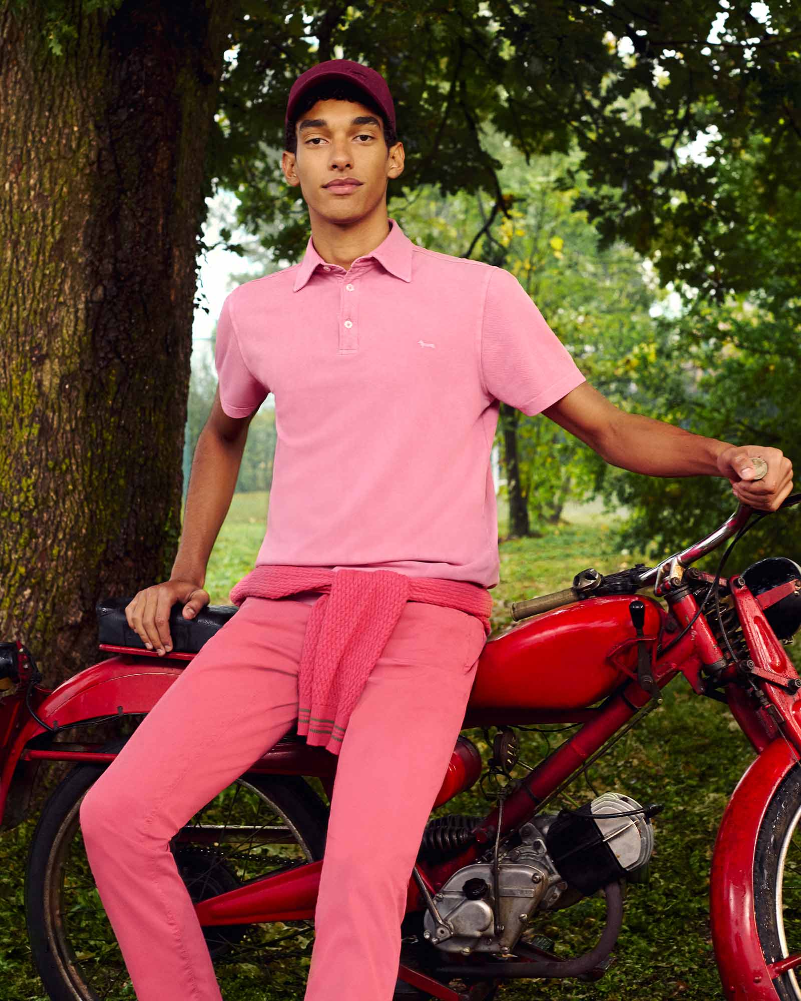 Young man in a monochrome pink outfit, sitting on a vintage red motorcycle in a green park — sporty and confident casualwear with a retro vibe.