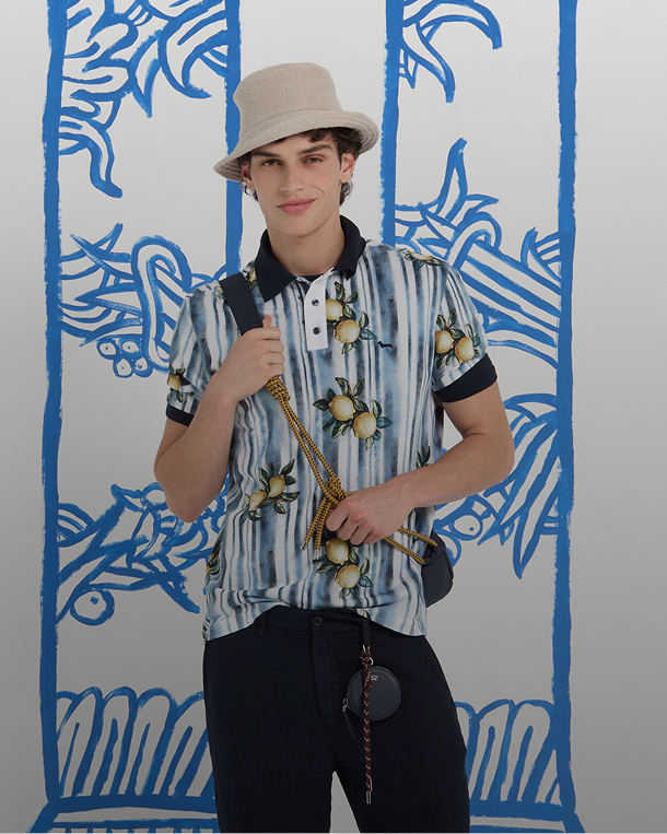 un uomo con cappello alla pescatora, polo a righe con stampa di limoni e pantaloni scuri posa davanti a uno sfondo con colonne blu. in basso compare il testo i trend del momento e la scritta per lui.
