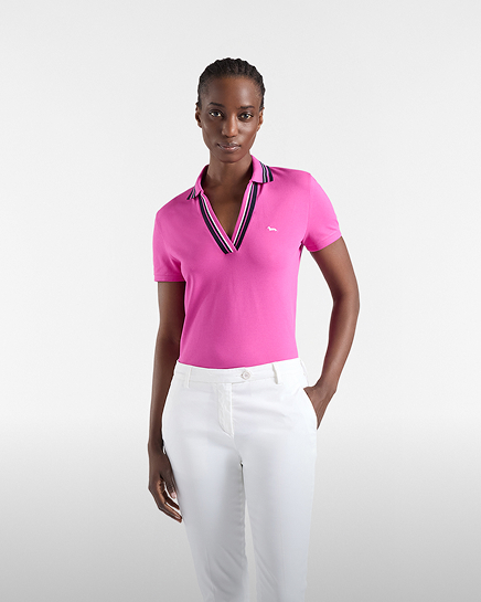 Promotional image focused on the women's polo category: a model poses facing forward against a light gray background wearing a bright pink short-sleeved polo shirt. the polo features an open collar detailed with contrasting white and dark blue striped borders, and the white dachshund logo embroidered on the chest. the model completes the look with straight-leg white trousers, tucking her left hand into the pocket.