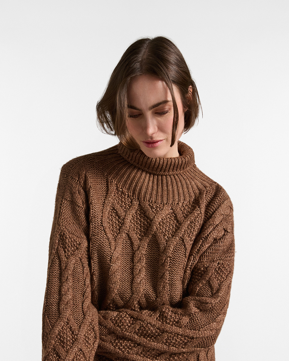 Close-up of a female model wearing a brown turtleneck sweater with cable-knit and textured patterns.