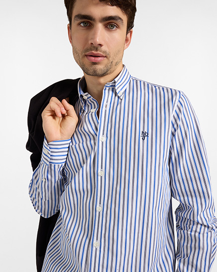 For the HB Jeans shirts category, a model wears a shirt with white, blue, and grey vertical stripes, featuring a button-down collar and a embroidered chest logo, while holding a black jacket over his shoulder against a white background.
