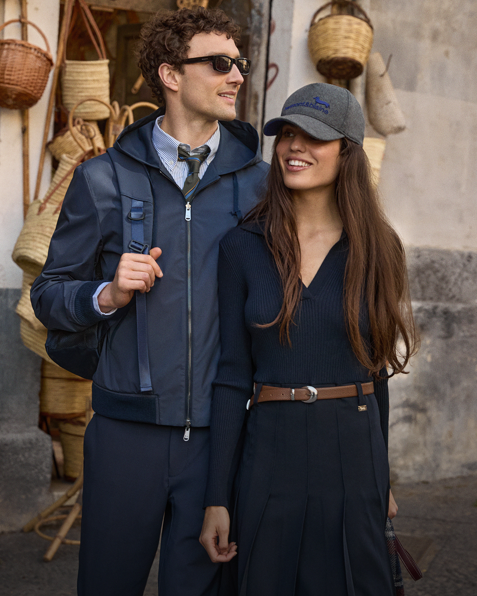 Promotional image for the Fall/Winter 2026 collection featuring a man and a woman side by side in front of a craft shop with hanging straw baskets; the man wears a dark blue hooded windbreaker, striped shirt, tie, and sunglasses, while the woman wears a dark blue ribbed sweater, brown leather belt, and a grey baseball cap with an embroidered logo.