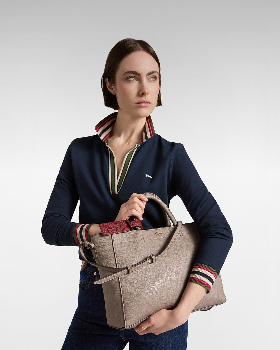 A woman with short brown hair wearing a navy blue long-sleeved polo shirt with a V-neck and striped trim, holding a taupe pebbled leather tote bag with top handles and a shoulder strap.