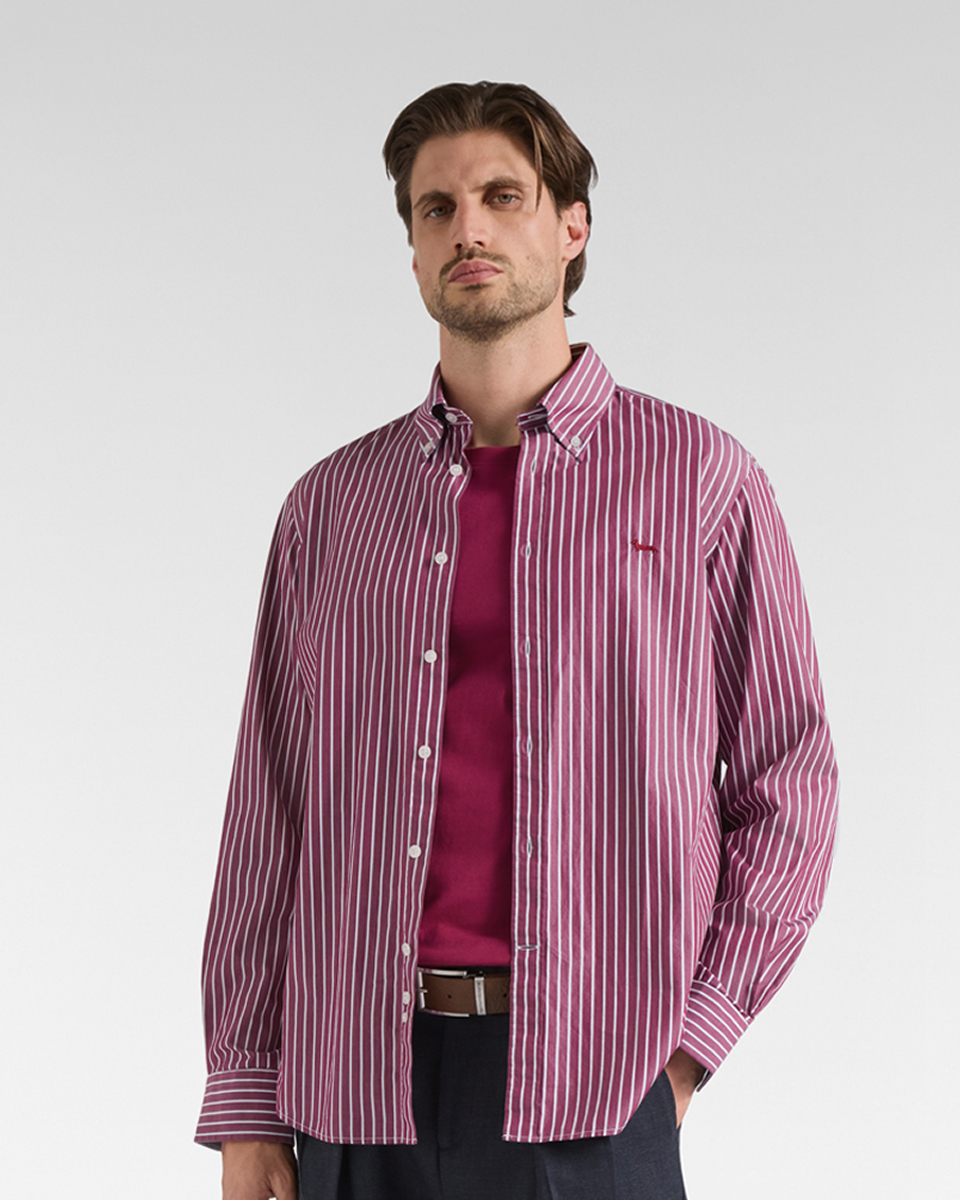 A brown-haired man with a short beard wearing an unbuttoned long-sleeved shirt with burgundy and white vertical stripes over a burgundy t-shirt and dark trousers with a belt.
