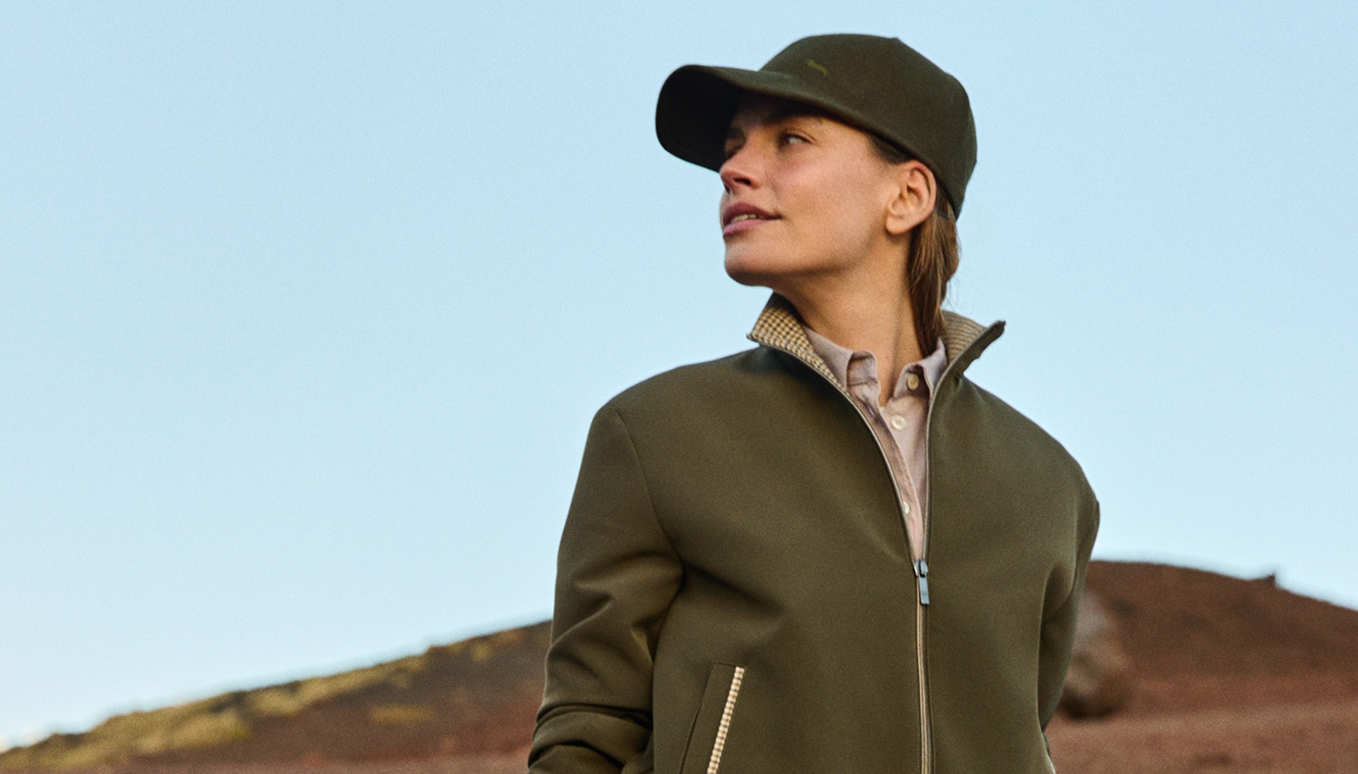 Female model wearing an olive green jacket, matching cap, patterned trousers, and a sweater tied around her waist, holding a small faux-shearling handbag against a rocky volcanic background.