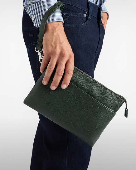 A close-up shot of a person wearing a striped shirt and dark jeans holding a dark green leather clutch bag with a wrist strap and zip closure.