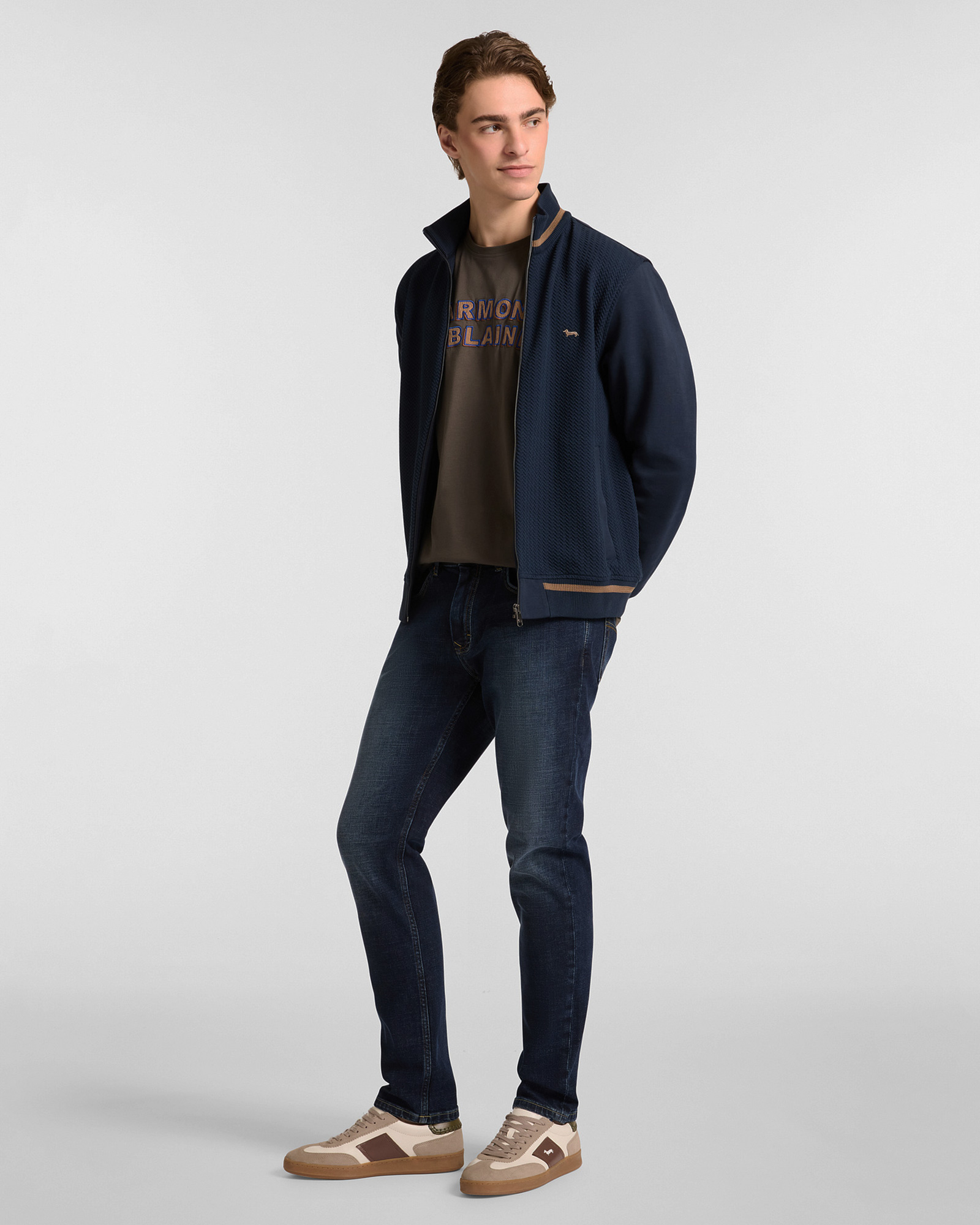 Uomo con giacca sportiva blu, t-shirt logata e jeans scuri con sneakers