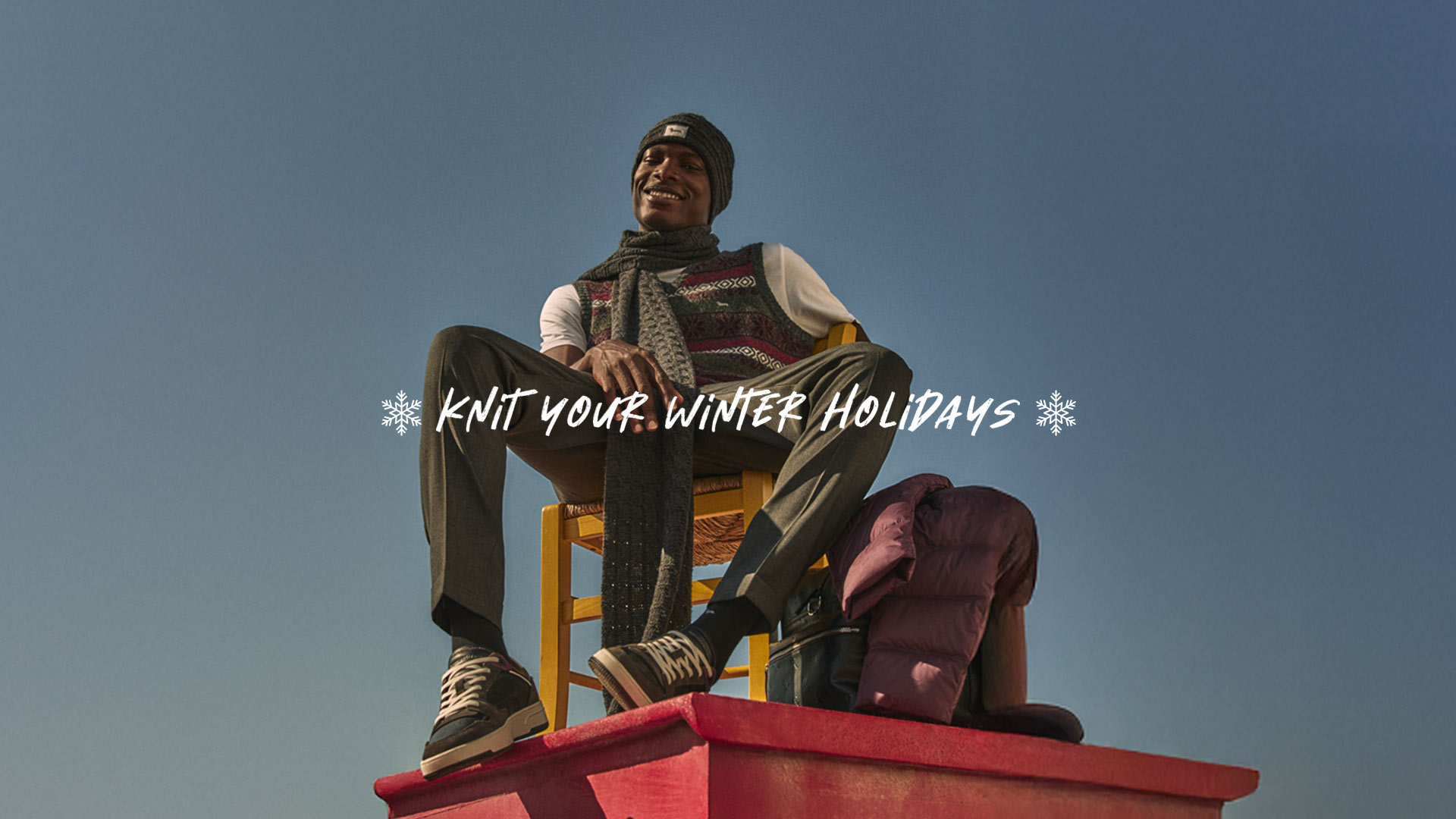 Man sitting outdoors wearing a knitted vest, grey scarf, beanie and tailored trousers.