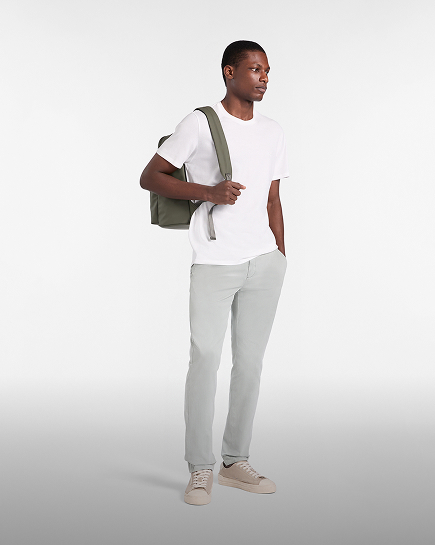 Full-length male model wearing a white crew-neck t-shirt and light grey trousers, carrying an olive green backpack over his shoulder and wearing beige sneakers, set against a gradient grey background.