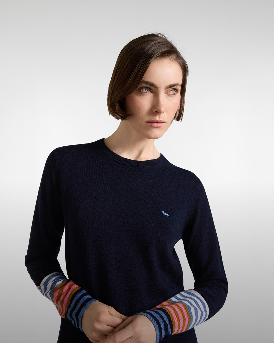 Woman with a bob haircut wearing a dark blue crewneck sweater with a light blue logo on the chest and multicoloured striped cuffs.