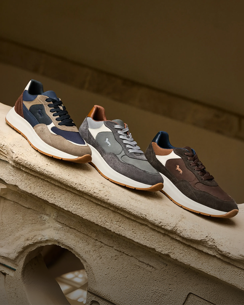 Tre paia di sneaker da uomo della collezione Autunno/Inverno 2026 disposte in fila su una balaustra in pietra decorata, realizzate in camoscio e tessuto tecnico multi-materiale nei toni del blu, grigio, marrone e beige, tutte caratterizzate dal logo del bassotto ricamato sul lato e da una suola spessa bianca con battistrada in gomma.