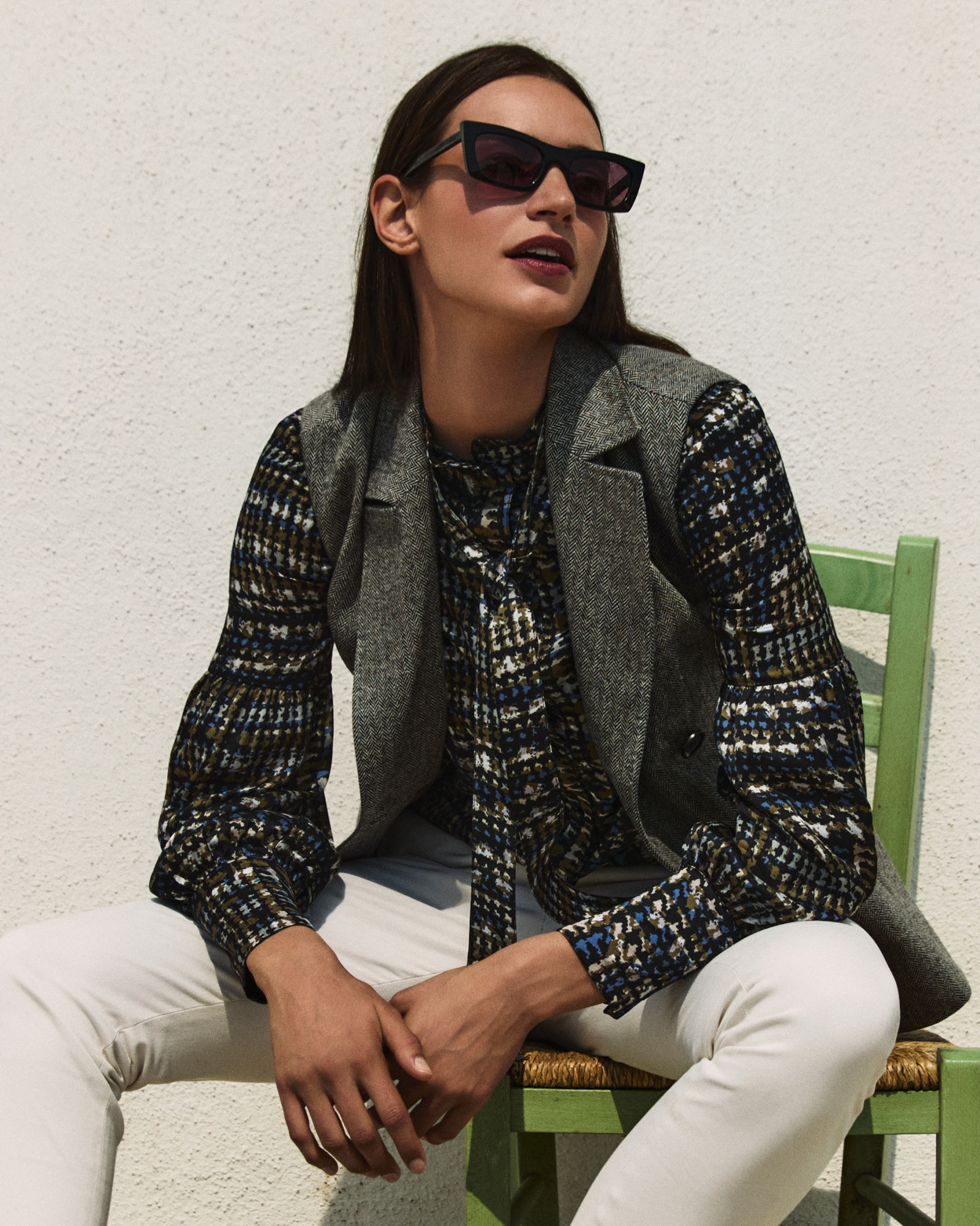 Woman wearing a grey vest, patterned shirt, and light trousers.