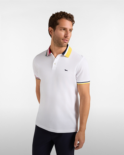 A man with curly brown hair and a short beard wearing a white short-sleeved polo shirt with a multicoloured collar and cuff trim, a small embroidered logo on the chest, and dark trousers.