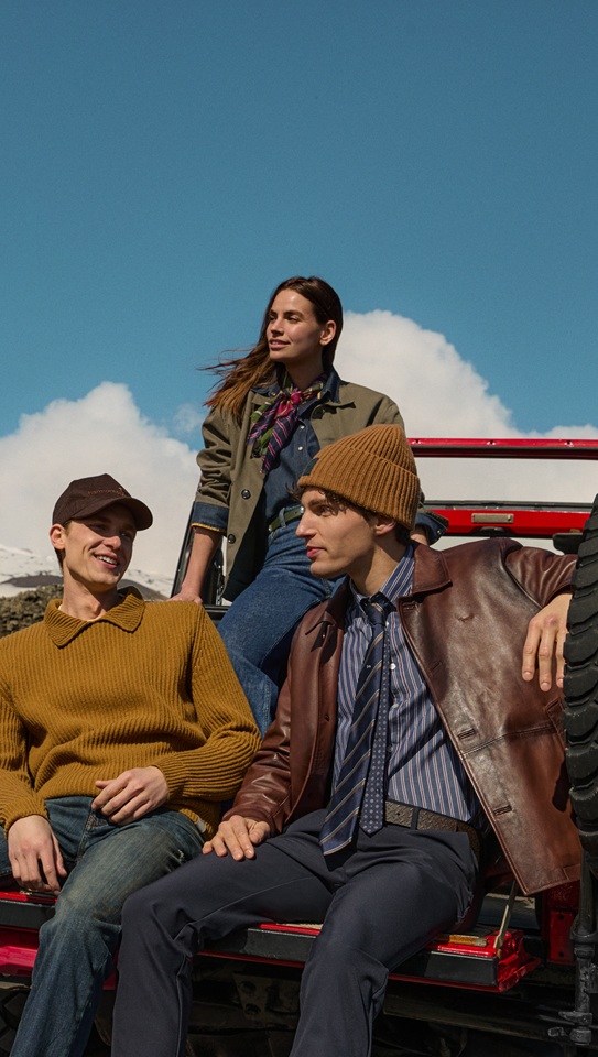 Three models in casual daywear and outerwear sitting on the back of a red off-road vehicle against a snowy mountain landscape.