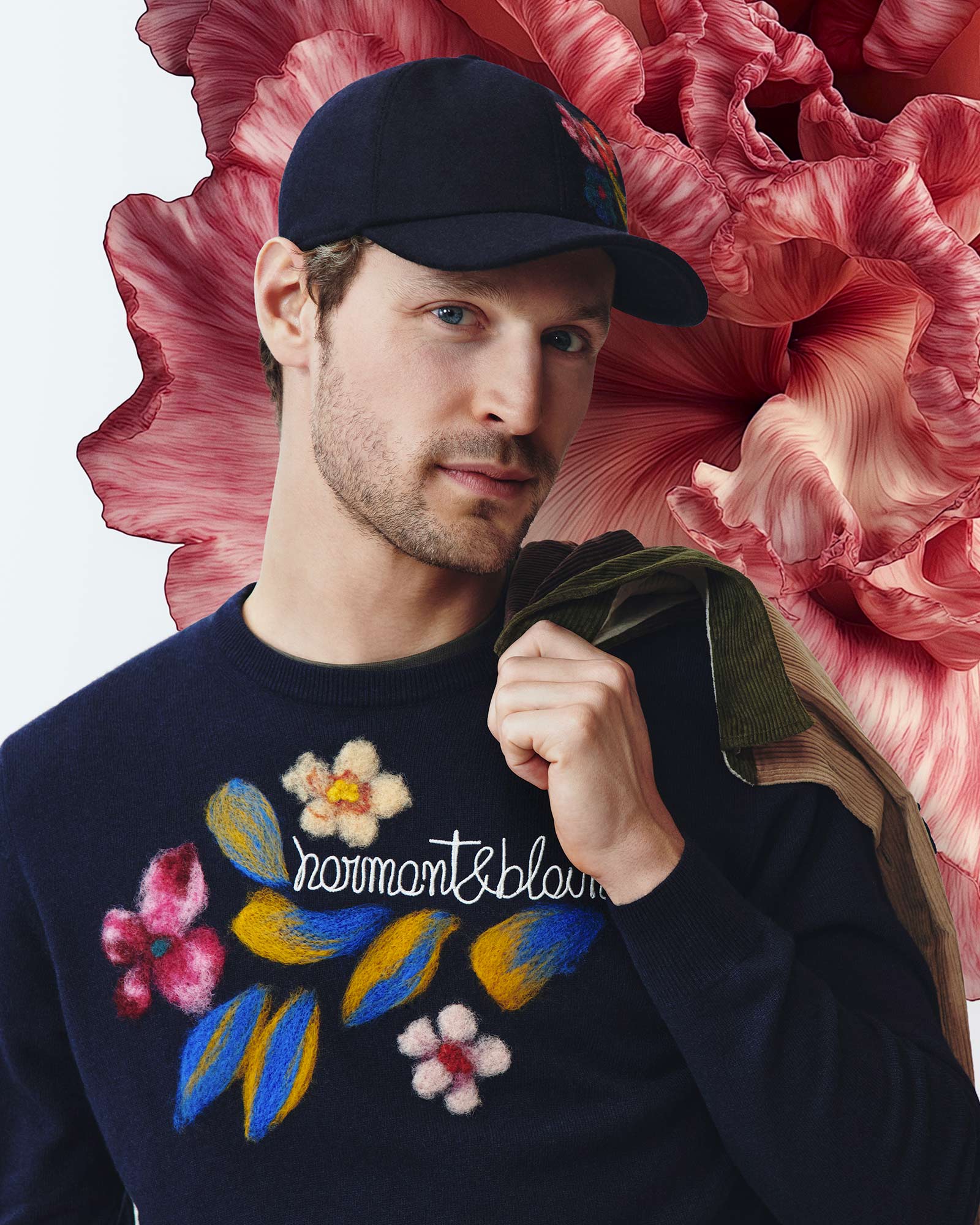 Man wearing a blue sweater with colorful floral decorations and a blue cap in front of a red flower.