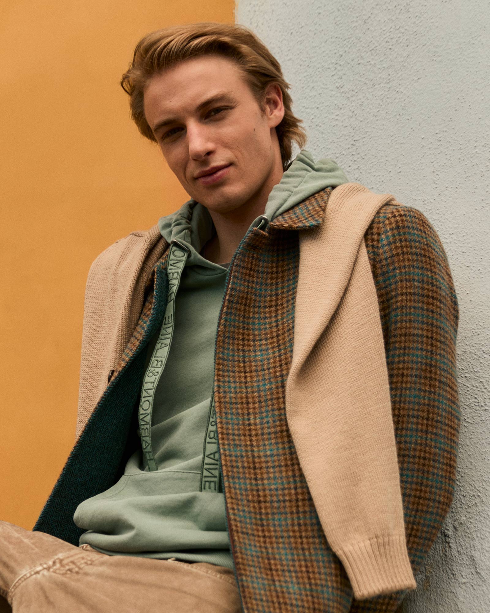 Male model wearing a brown and green checked coat with a sage green hoodie.