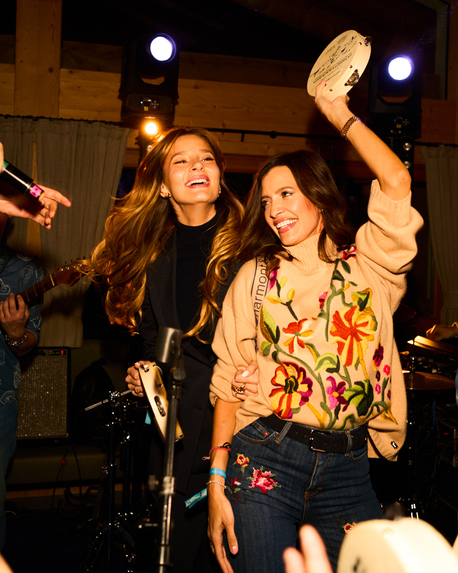 Due donne che ballano con tamburelli, indossando maglione floreale e look scuri.