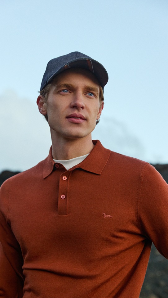 Male model wearing a rust-colored long-sleeve polo shirt and a dark baseball cap, posing outdoors in front of a hilly landscape under a cloudy sky.