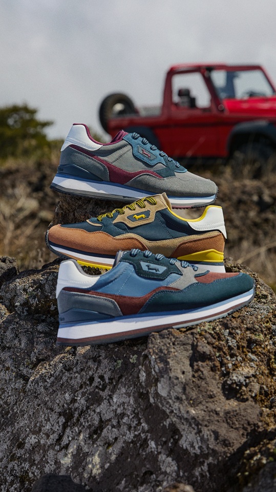 Three pairs of multicolored sneakers stacked on top of a rock, with a blurred red off-road vehicle in the background within a natural landscape.