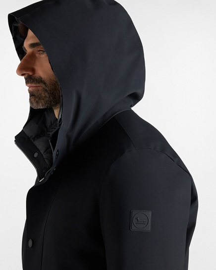 This promo image features a close-up profile shot of a man wearing a dark hooded jacket with a padded inner bib and a tonal Dachshund logo patch on the sleeve. The primary focus of the visual is to highlight Harmont & Blaine men's clothing, showcasing the modern design, functional details, and technical quality of the outerwear collection.