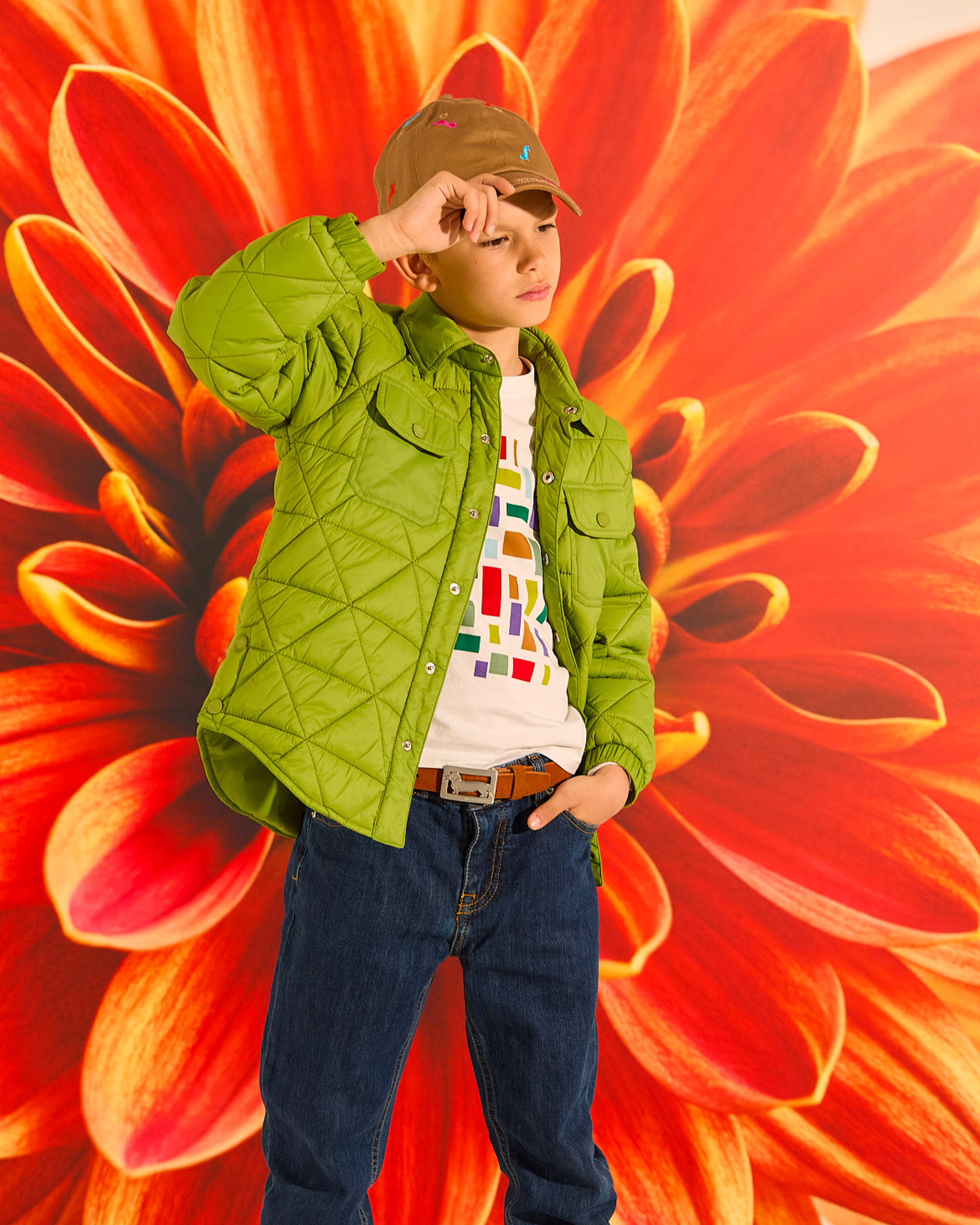 Boy in green quilted jacket, printed T-shirt, jeans and beige cap